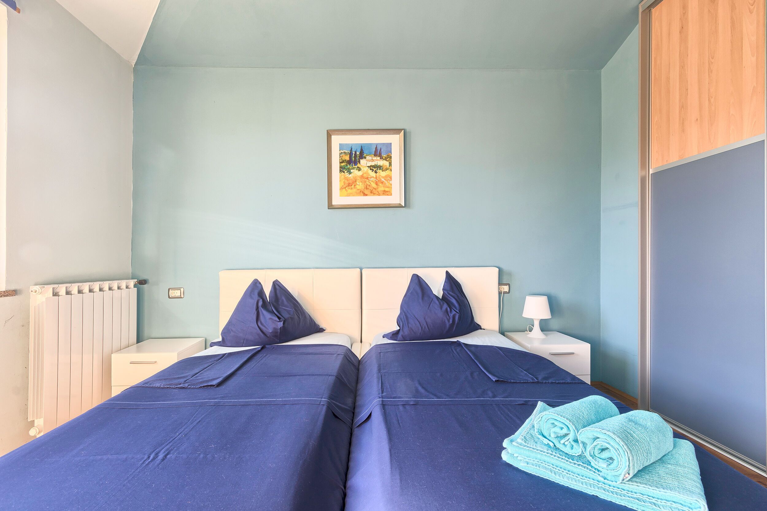 A room with two beds, a nightstand with a lamp, a radiator, and a framed picture on the wall.