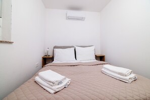 Una cama individual con cubrecama acolchada, dos almohadas, una mesita de noche con lámpara y un aire acondicionado de pared.