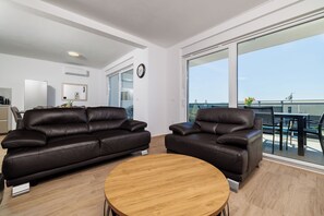 Un salón moderno con dos sofás de cuero marrón, una mesa de centro de madera, un ventanal con vista al mar y una cocina al fondo.