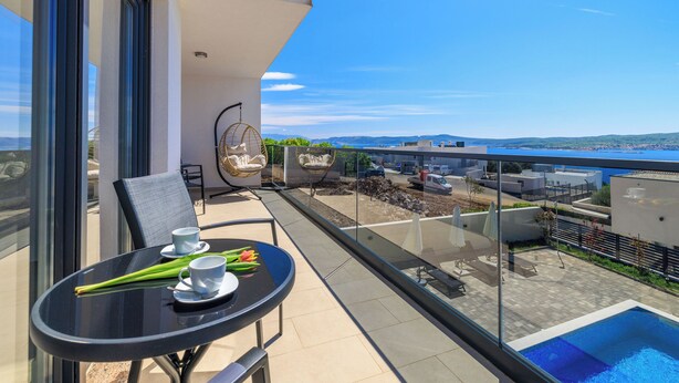 En balkong med ett bord dukat för två personer, en pool och utsikt över havet och byggnaderna.