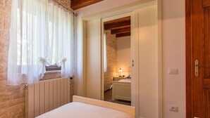 A bedroom with a bed, a window with sheer curtains, a wooden door, and a view into an adjacent room with a sink and a lamp. - Villa Casa Mia (Barban)