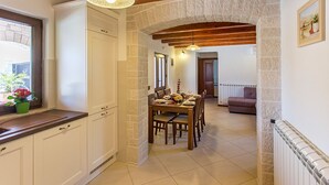 A kitchen with white cabinets, a dining table set for four, and a stone archway leading to a living area with a sofa. - Villa Casa Mia (Barban)