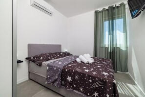 A single bed with a headboard, a bedside table, a lamp, a window with curtains, and an air conditioning unit. - Villa Hermosa (Vinodol)