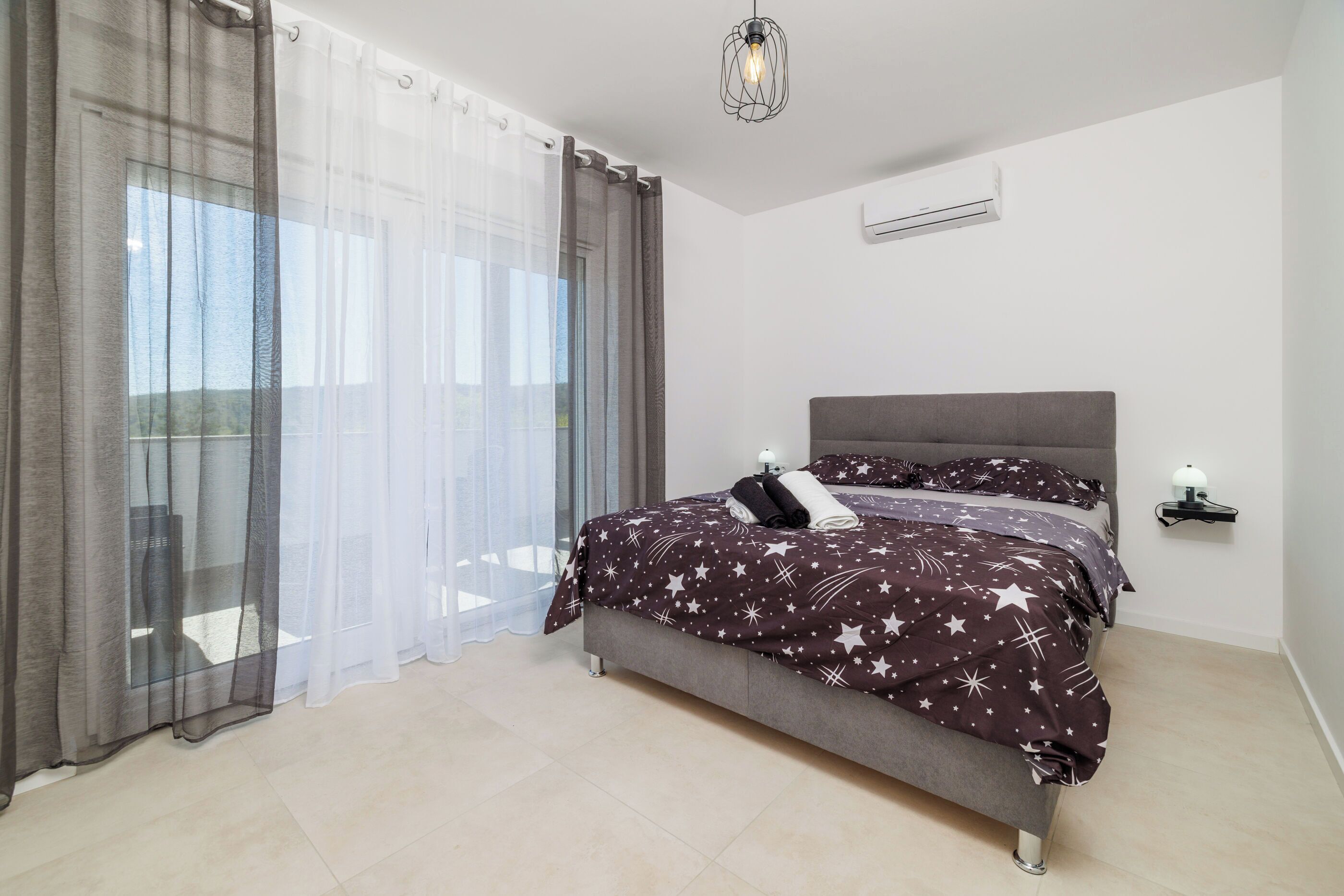 A bedroom with a bed, curtains, a bedside table, and a hanging light fixture.
