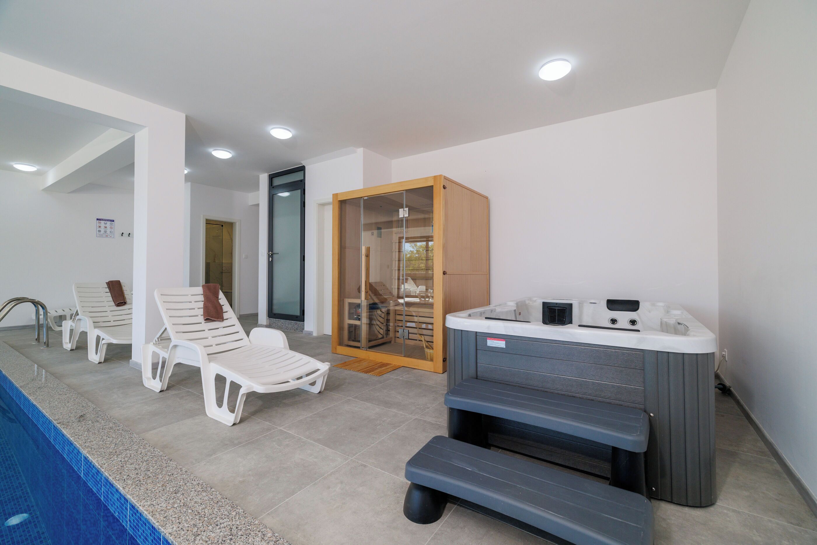 A modern indoor pool area with lounge chairs, a hot tub, and a glass-enclosed sauna.