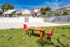Ein grüner Außenbereich mit Tisch und Stühlen, einer weißen Mauer und einem Gebäude mit rotem Dach im Hintergrund.