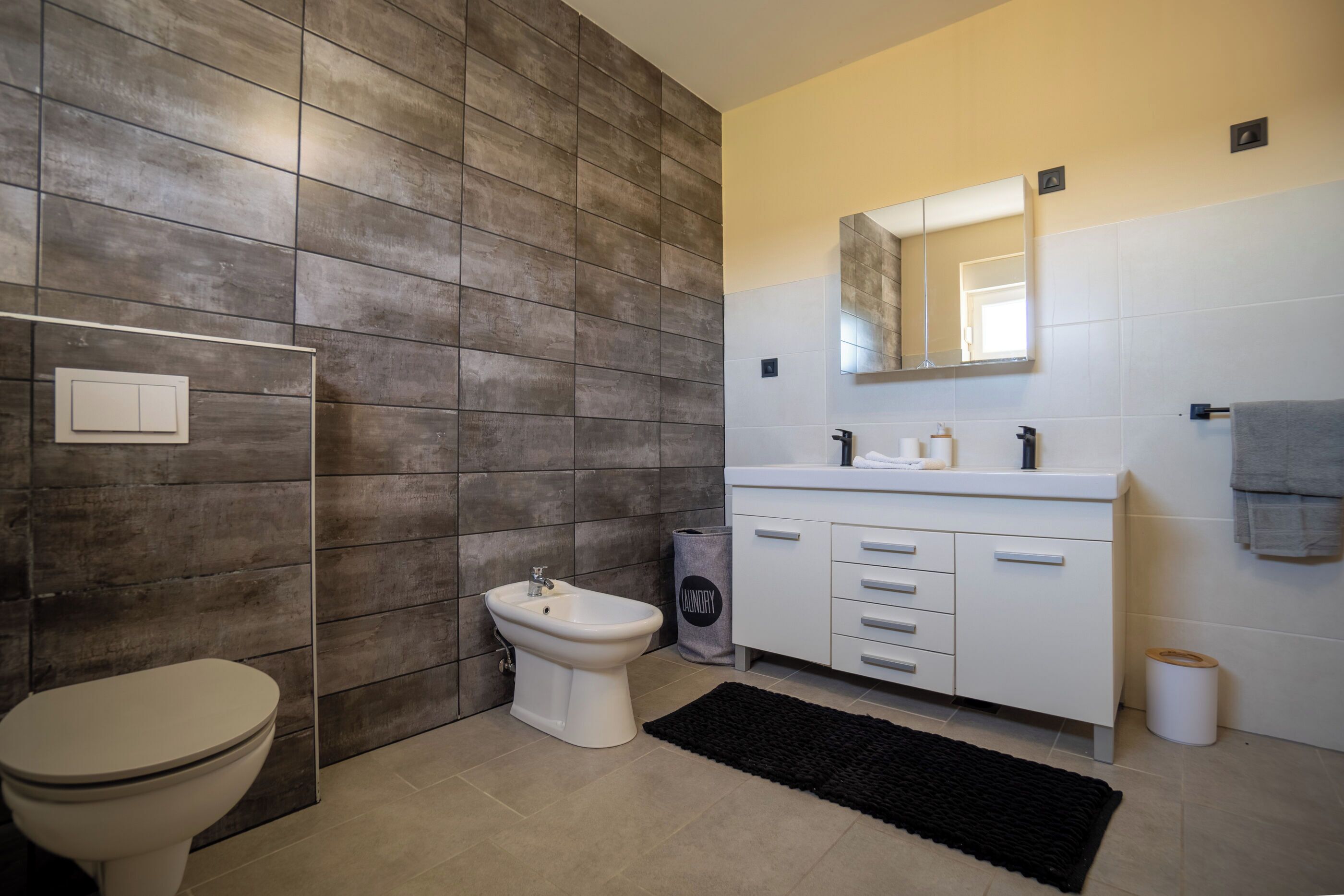 Ein modernes Badezimmer mit integriertem Bidet, Toilette, weißem Waschtisch mit Spiegel und einem schwarzen Teppich.