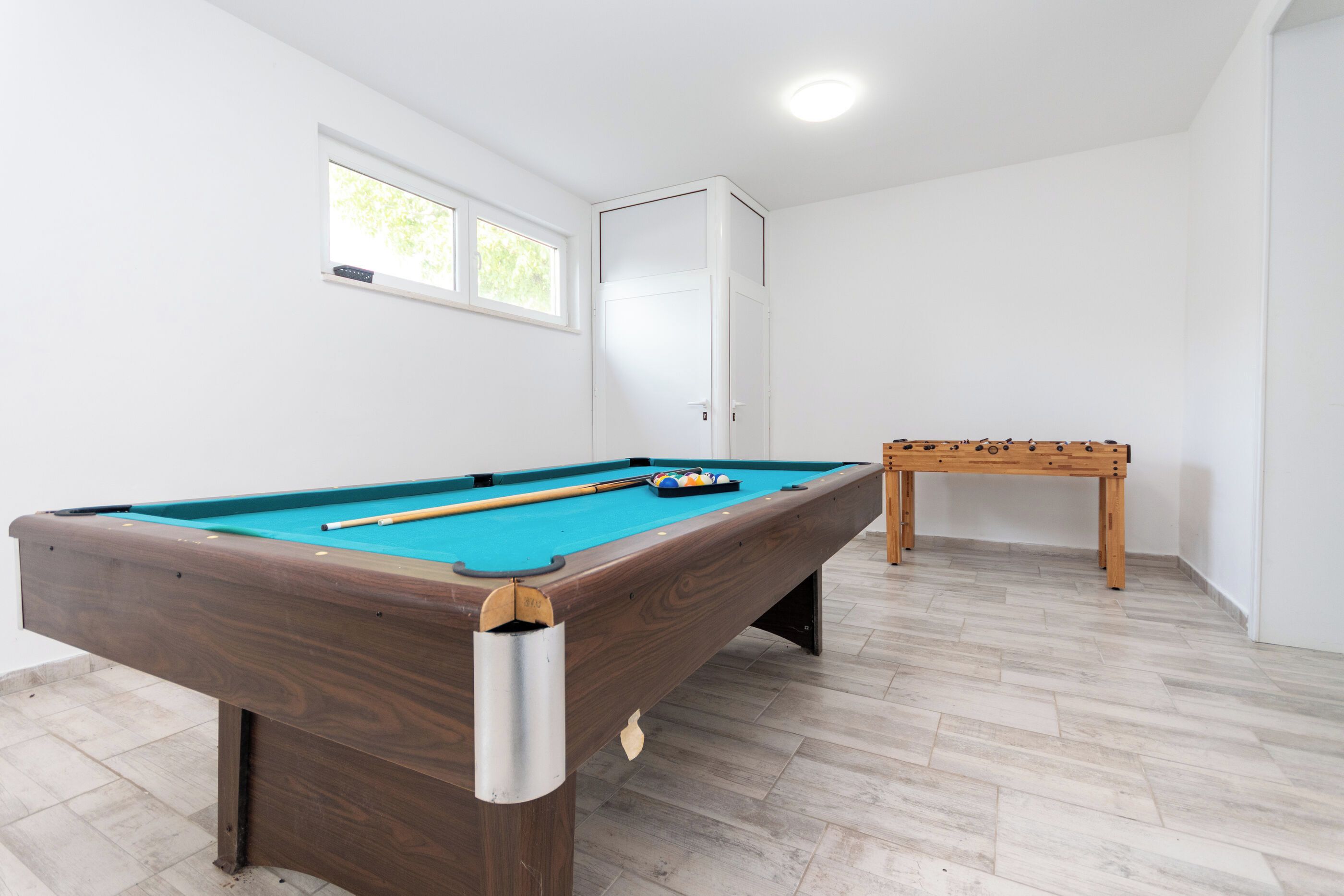 Een pooltafel met houten frame en blauw vilt, een tafelvoetbalspel, en een raam met natuurlijk licht.
