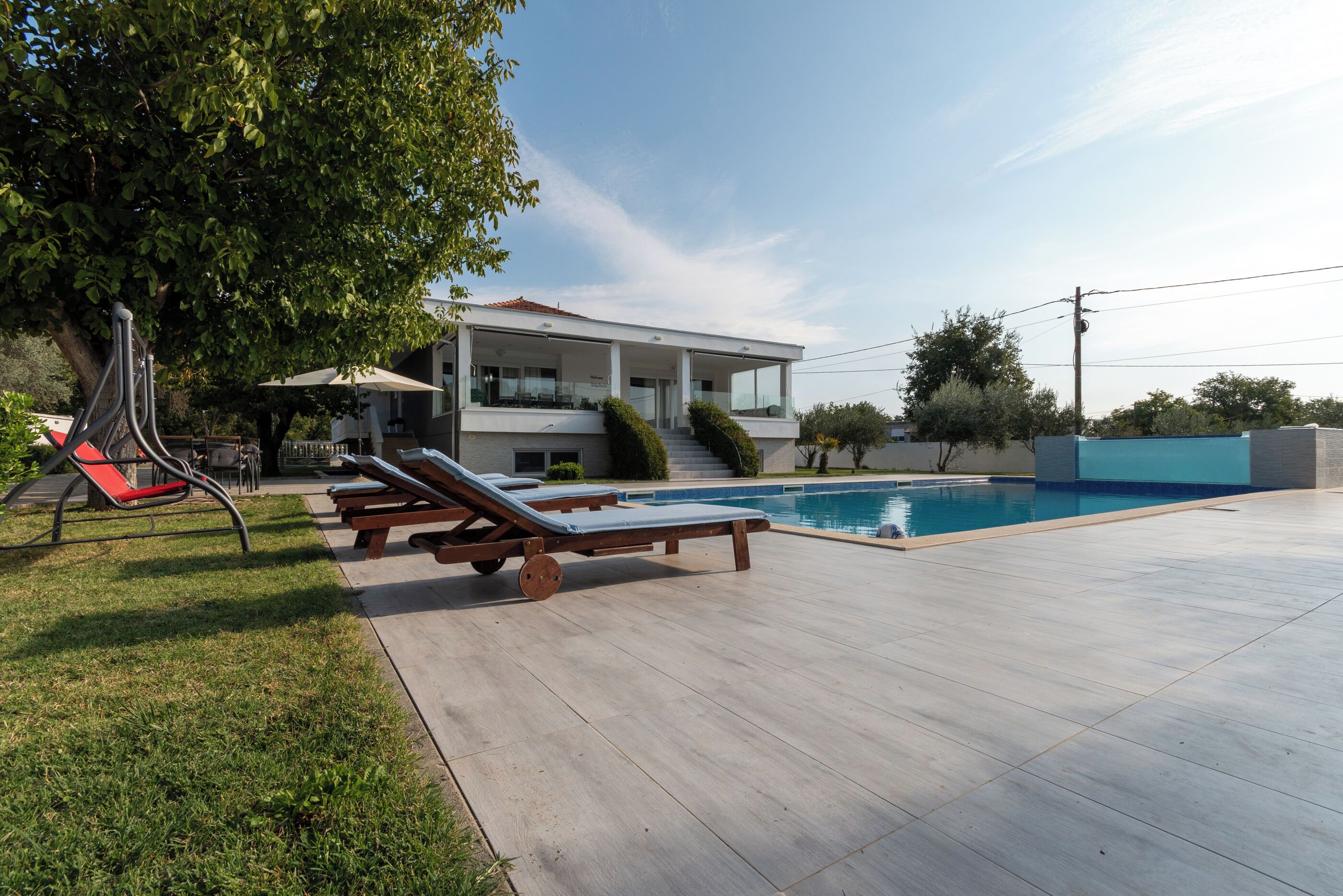 Una casa moderna amb una piscina, mobles d'exterior i un arbre que fa ombra.