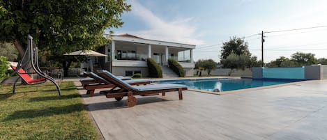 Una casa moderna amb una piscina, mobles d'exterior i un arbre que fa ombra.