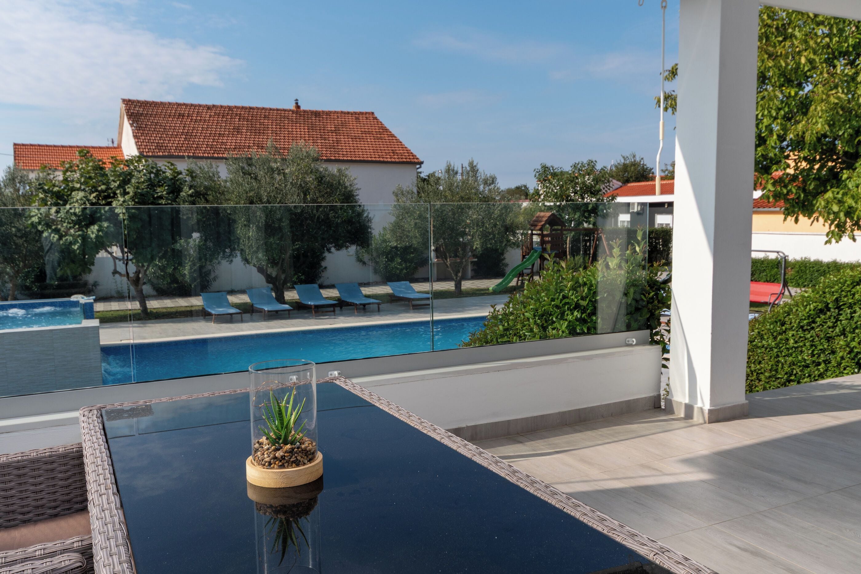 A poolside area with a glass table, a potted plant, and a view of a building with a red-tiled roof.