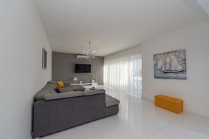 Ein modernes Wohnzimmer mit grauem Sofa, einem an der Wand montierten Fernseher, einer Chandelieber und einem Gemälde eines Segelboots.