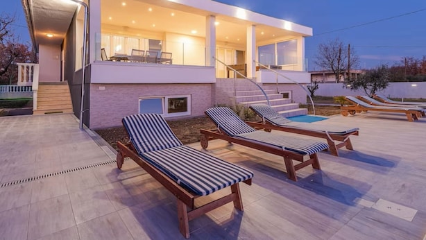 Een modern huis met een zwembad, ligstoelen en een overdekt terras.