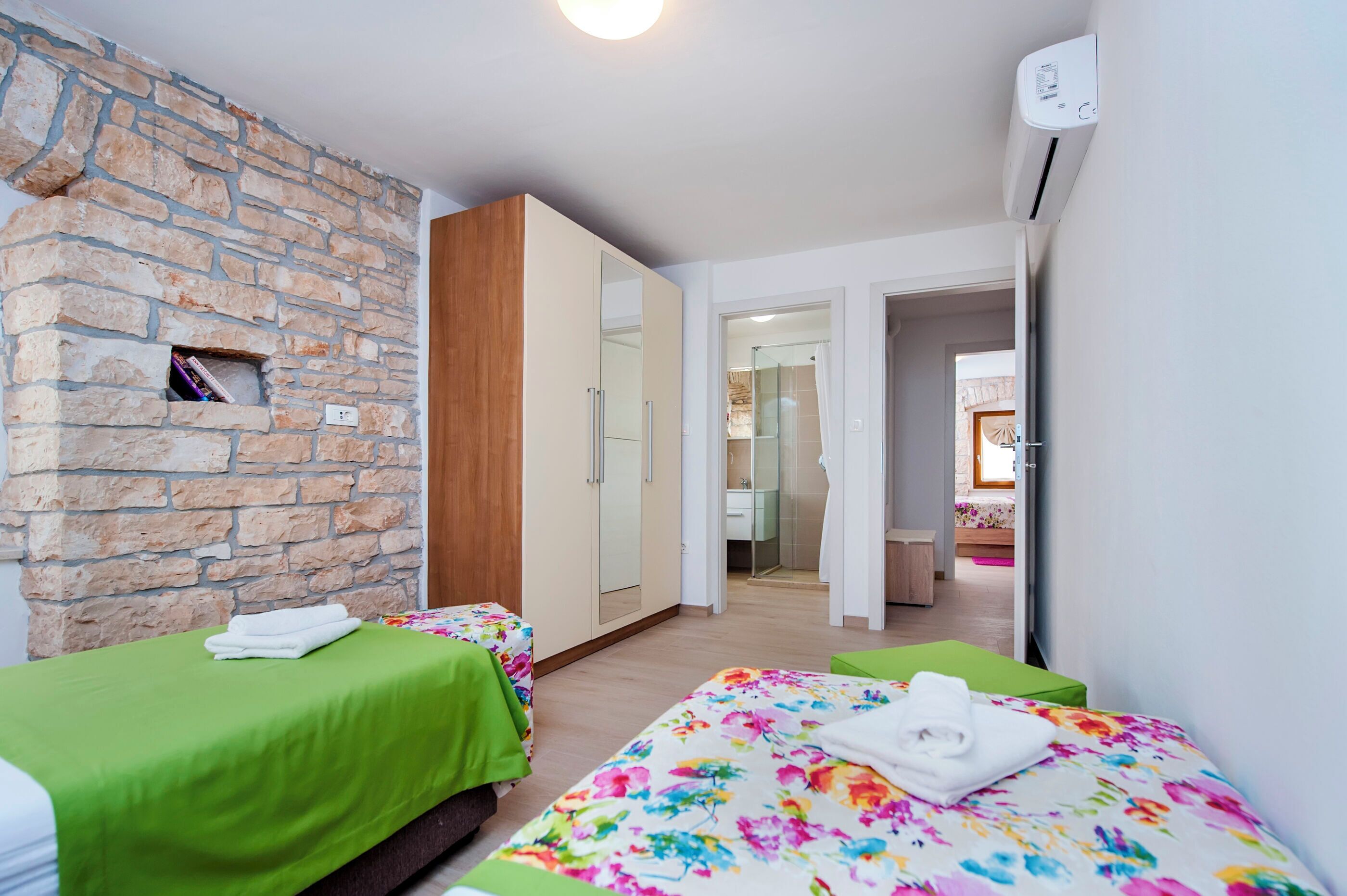 Une pièce avec un mur en pierre, une armoire en bois, un lit recouvert d’une courtepointe à motifs floraux et une autre courtepointe verte.