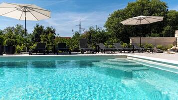 Ein Swimmingpool mit Liegestühlen und Sonnenschirmen, umgeben von Bäumen und blauem Himmel.