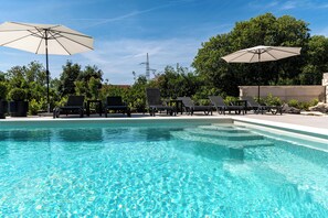 Ein Swimmingpool mit Liegestühlen und Sonnenschirmen, umgeben von Bäumen und blauem Himmel.