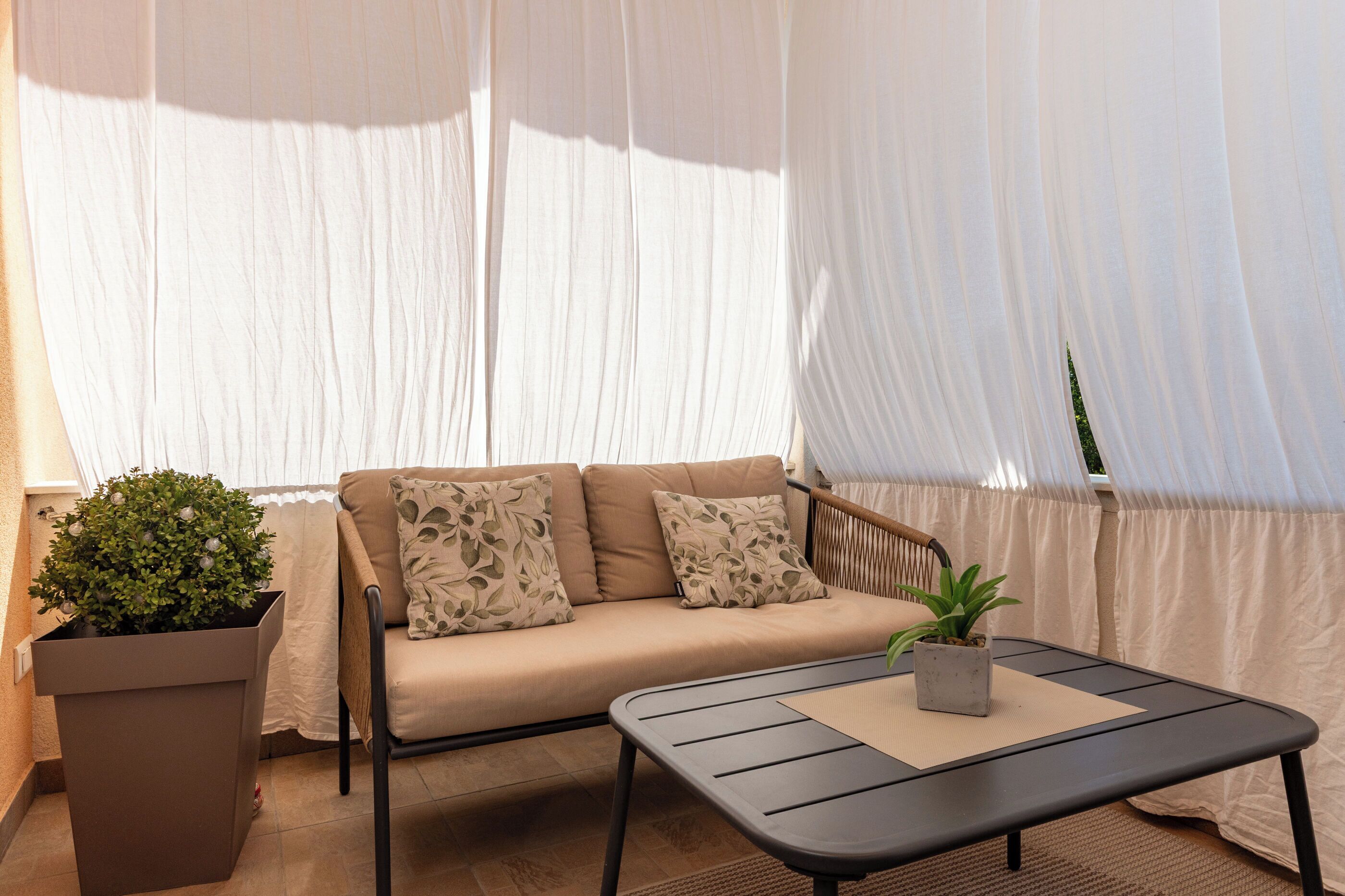 Een beige bank met decoratieve kussens, een klein tafeltje met een plant erop, en een potplant naast de bank.