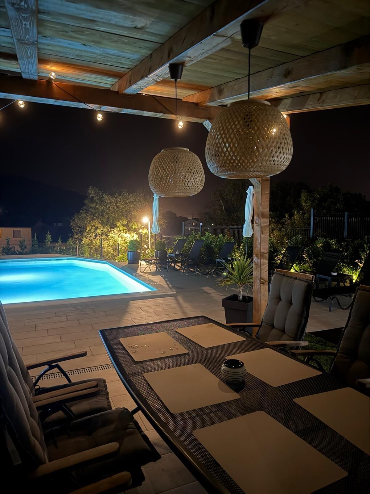 Un espace extérieur avec une piscine, des chaises longues et des tables, entouré d’une terrasse en bois et surplombé d’une pergola agrémentée de guirlandes lumineuses.