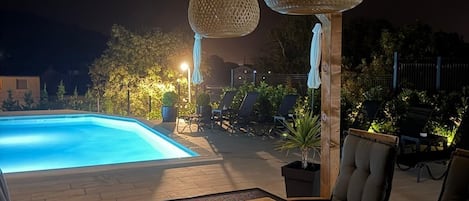 An outdoor pool area with lounge chairs and tables, surrounded by a wooden deck and under a pergola with hanging lights.