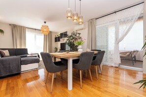 A modern living room with a dining table, grey chairs, a sofa, and a large window with curtains.