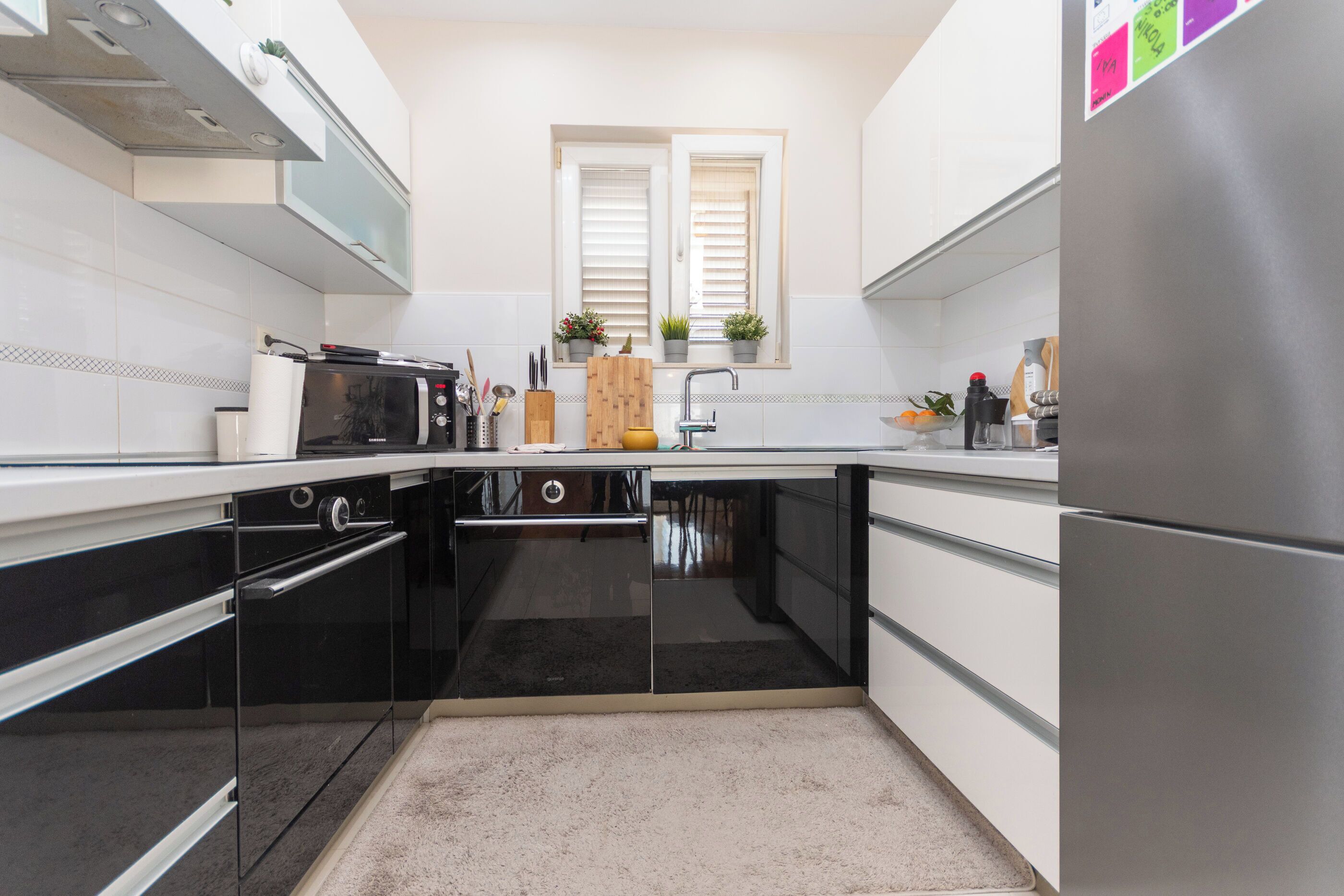 Una cocina moderna con electrodomésticos negros, armarios blancos, un ventanal con persianas y una pequeña planta en el alféizar.