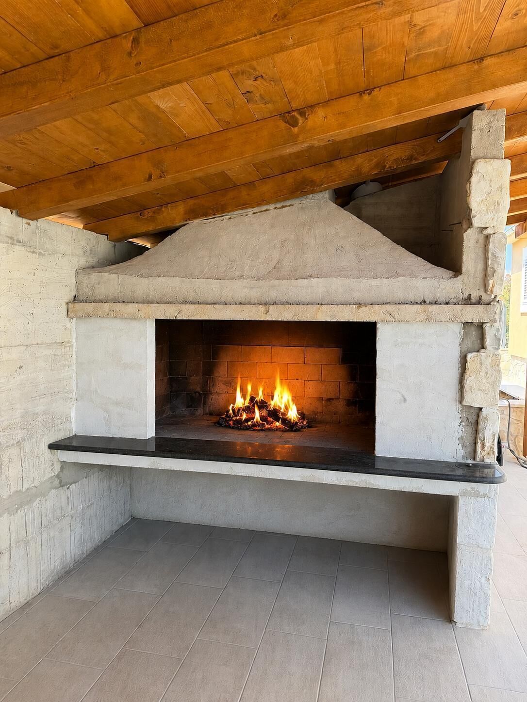 A stone-built outdoor fireplace with a fire burning inside, under a wooden roof structure.