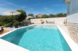 Ein rechteckiger Swimmingpool mit klarem, blauem Wasser, umgeben von einer Steinmauer und beschattet von Sonnenschirmen.
