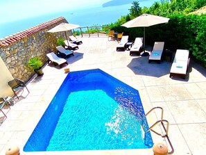 Une piscine entourée de murs en pierre, agrémentée de chaises longues et de parasols, et offrant une vue sur la mer.