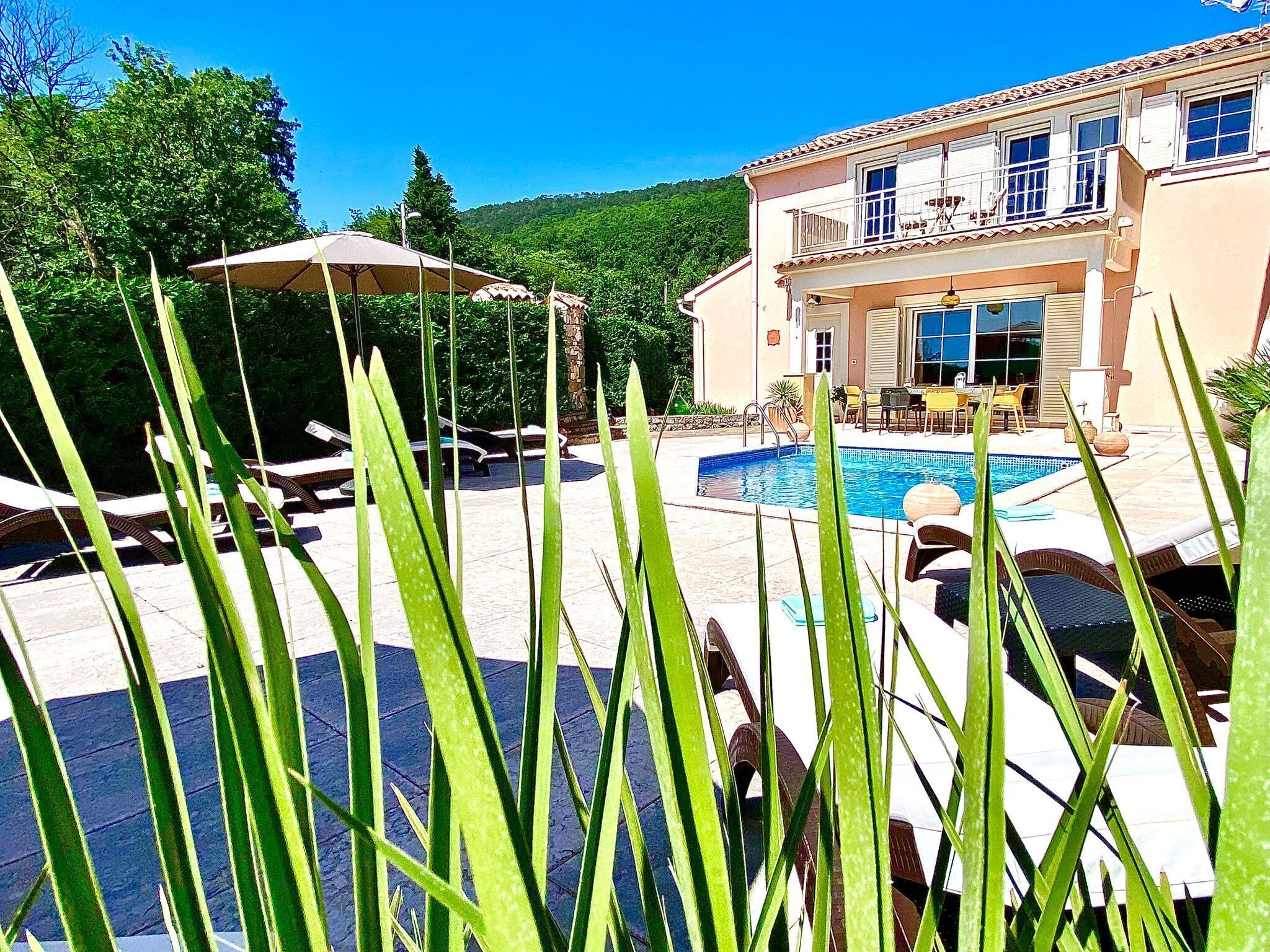 Un espace piscine avec des chaises longues et des parasols, un bâtiment à deux étages doté de balcons, et un paysage montagneux en arrière-plan.
