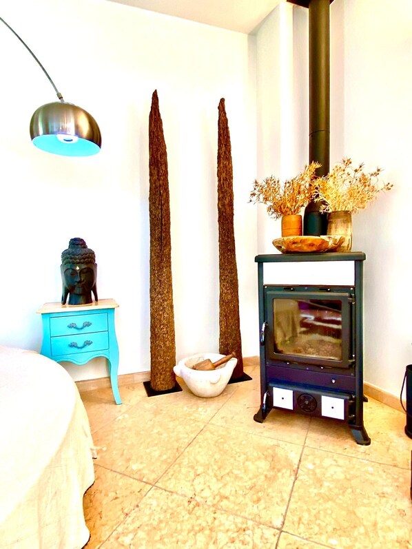Une chambre Ă coucher avec un lit, une table de chevet bleue, un bol en bois, un poĂŞle en pierre et une lampe suspendue.
