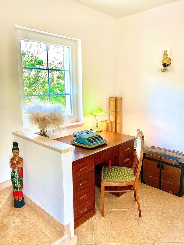 Ein Zimmer mit Schreibtisch, Stuhl, Fenster, Lampe und einer kleinen Statue.