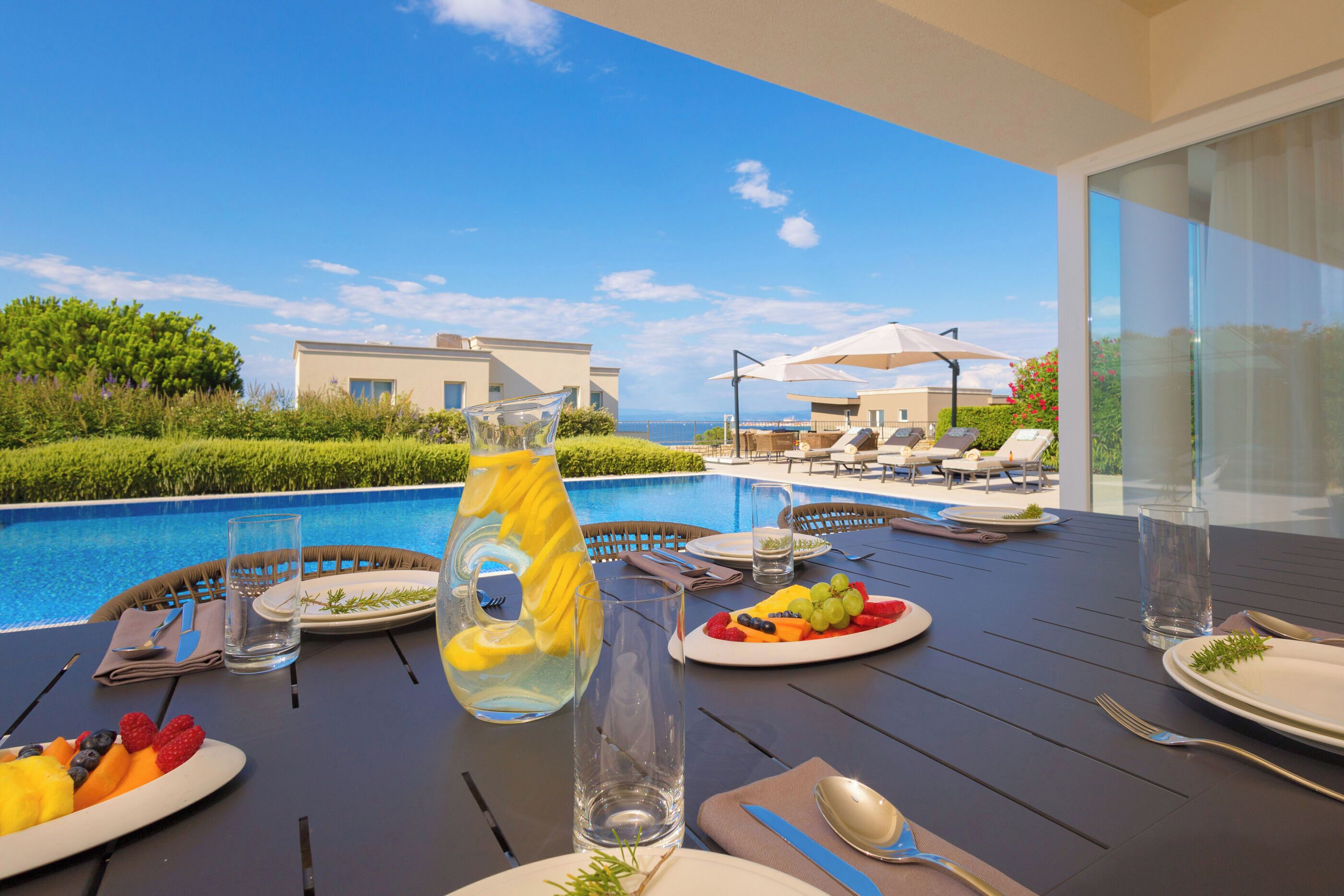 Una mesa de comedor con platos de fruta, una jarra y cubiertos, con vistas a una piscina y zona de estar.
