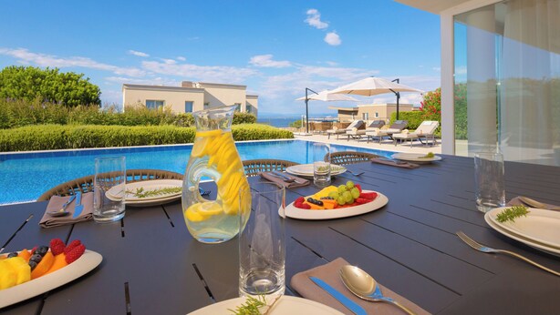 Una mesa de comedor con platos de fruta, una jarra y cubiertos, con vistas a una piscina y zona de estar.
