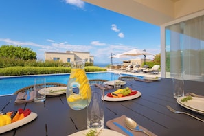 Mesa de jantar com pratos de fruta, jarra e talheres, com vista para uma piscina e área de estar.