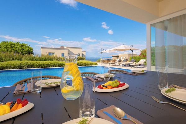 Una mesa de comedor con platos de fruta, una jarra y cubiertos, con vistas a una piscina y zona de estar.
