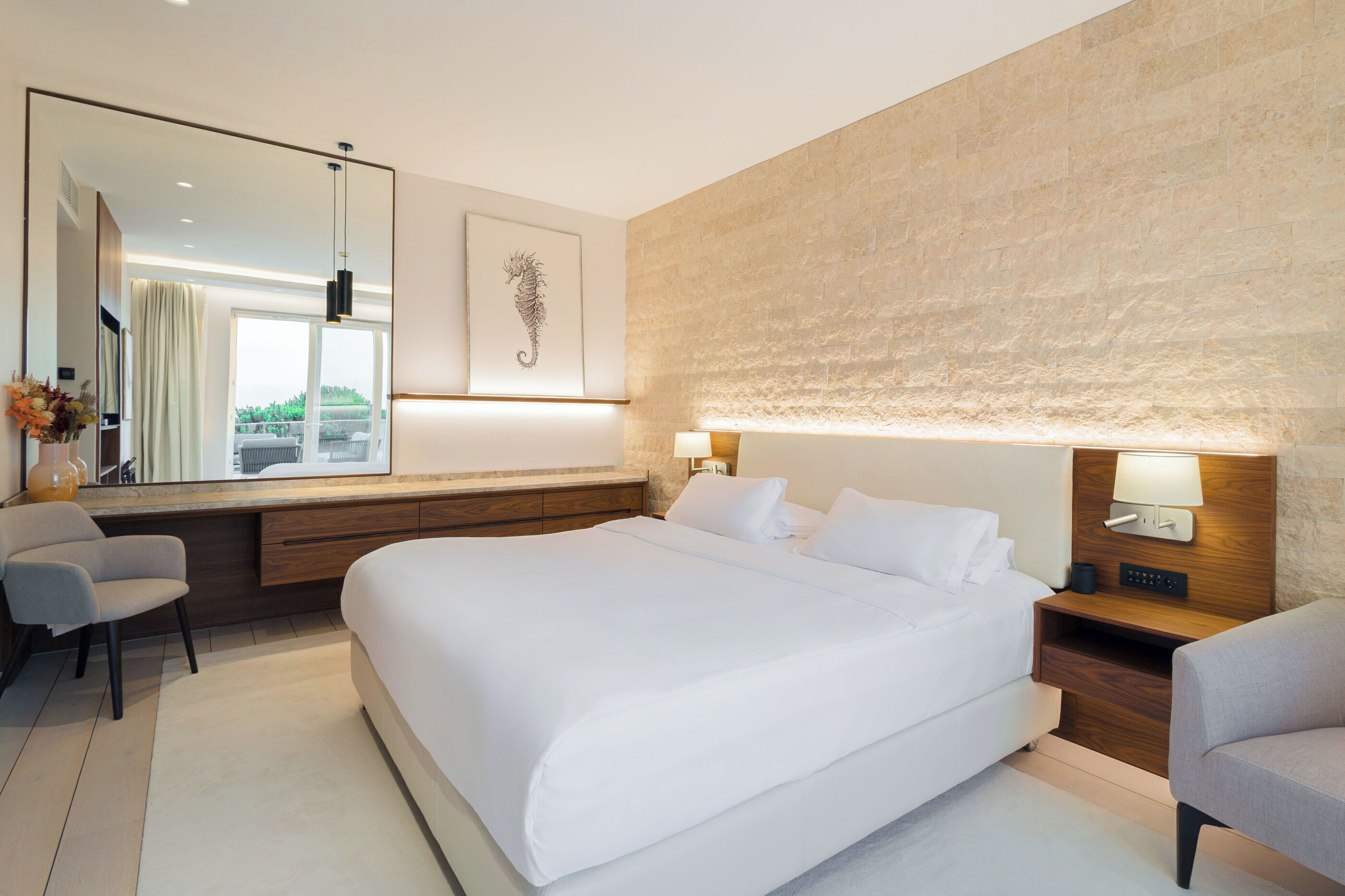 A modern hotel room with a large bed, a desk, a chair, a nightstand with a lamp, and a large mirror.
