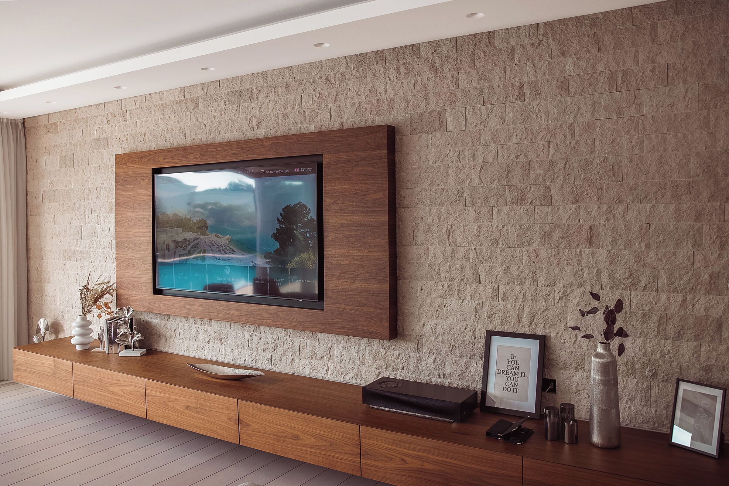 Uma sala de estar moderna com TV na parede, móvel de entretenimento em madeira e objetos decorativos.