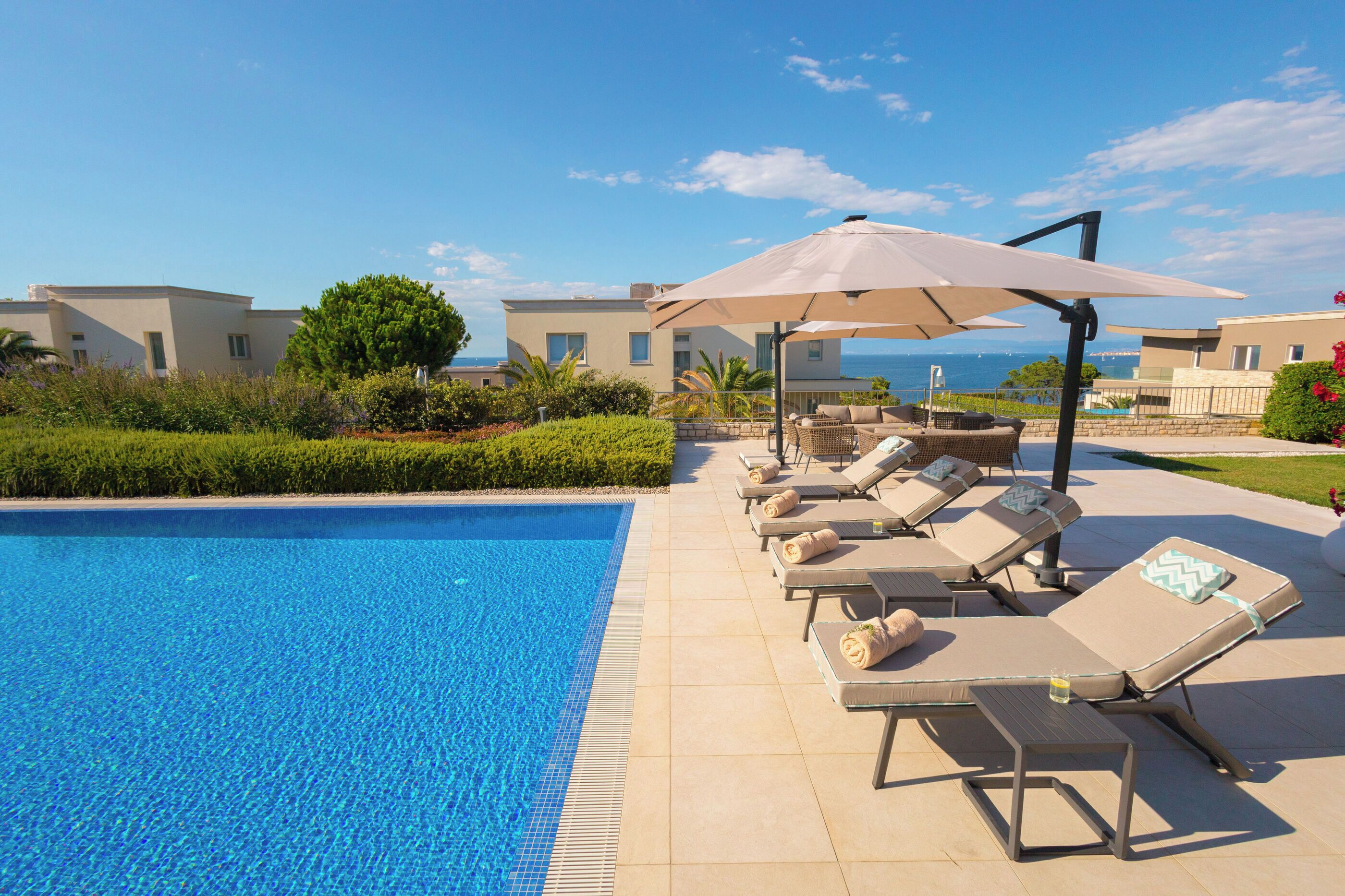 A swimming pool with lounge chairs and umbrellas, a clear sky, and buildings in the background.