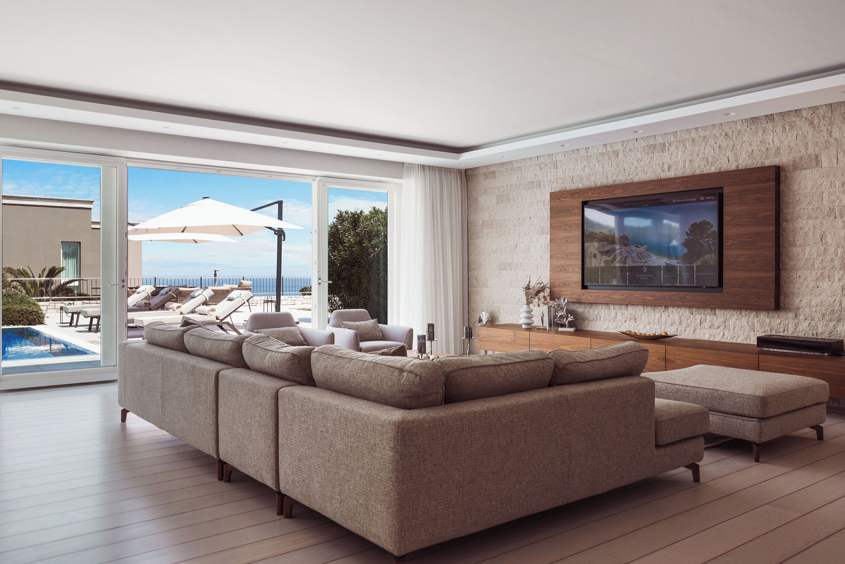 Uma sala de estar moderna com um amplo sofá modular, uma TV de tela plana fixada em uma parede texturizada, e vista para uma piscina e área de estar externa.
