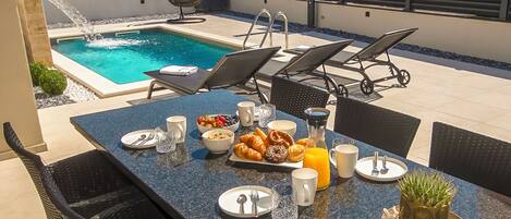 A poolside dining area with a table set for breakfast, including croissants, fruit, and juice.