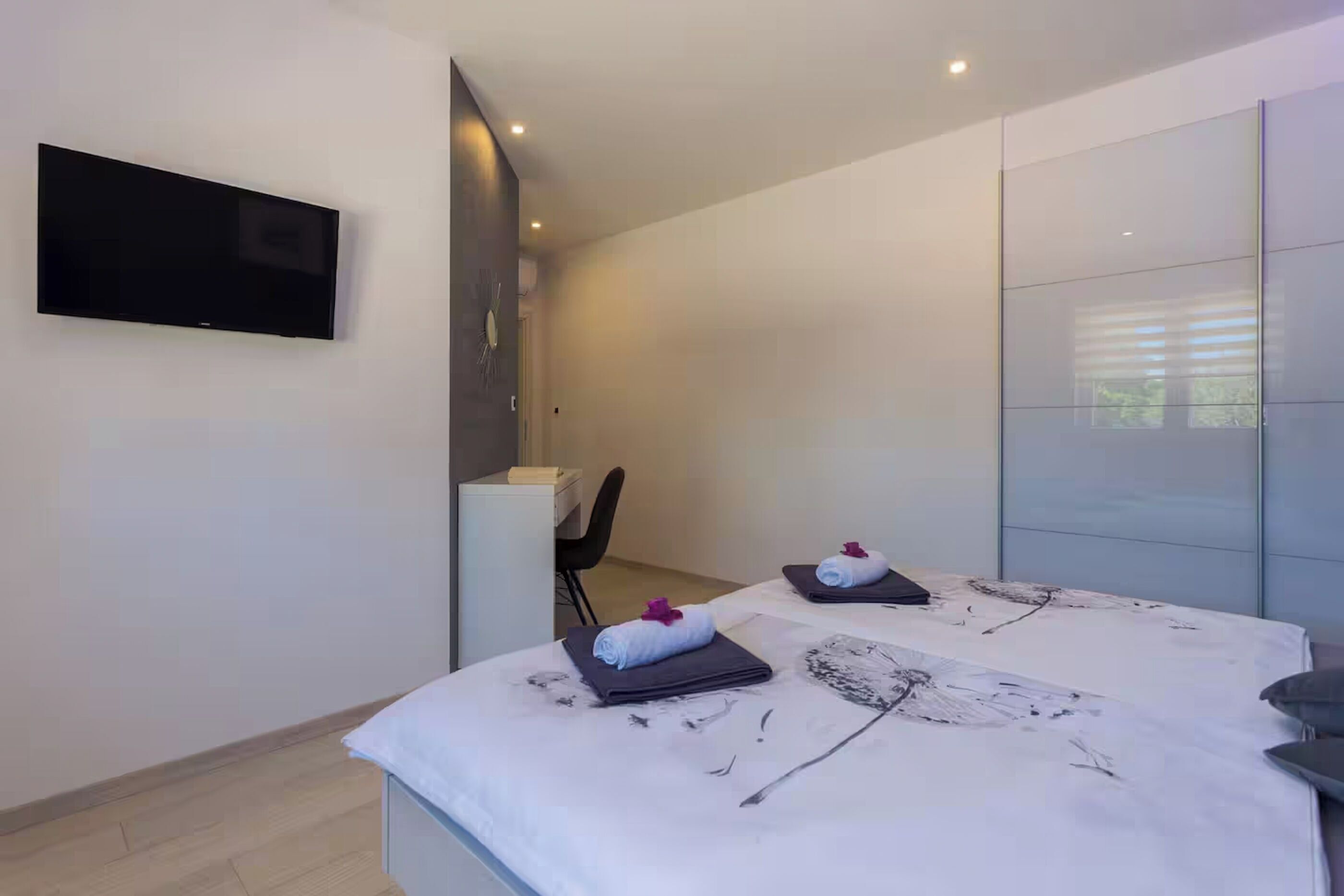 A modern hotel room with a flat-screen TV mounted on the wall, a bed with a white bedspread and a towel with a purple flower, and a large window with blinds.