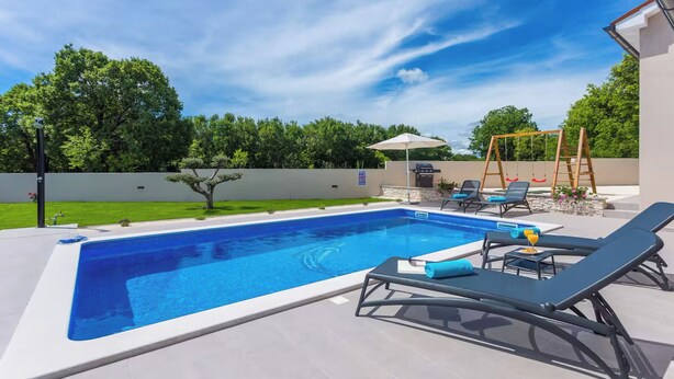 Una piscina rettangolare con superficie d'acqua cristallina blu, circondata da un solarium piastrellato. Si notano lettini prendisole e un tavolino con una bevanda, e in lontananza un'altalena in legno.