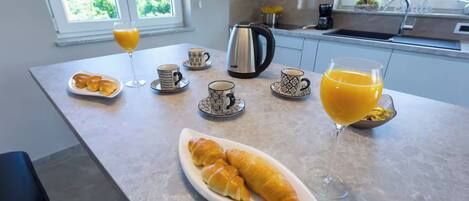 A kitchen counter with a croissant, coffee cups, a glass of orange juice, and a kettle.
