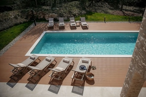 Une piscine rectangulaire, agrémentée de chaises longues et d’une table, entourée d’une terrasse en bois et d’un mur en pierre.