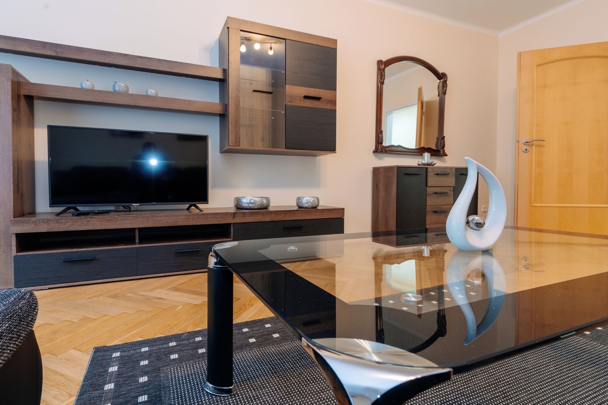 A modern living room with a flat-screen TV, wooden entertainment unit, glass coffee table, and a decorative vase.