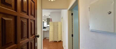 Un pasillo con una puerta de madera, una pared blanca con un letrero de emergencia, una cortina estampada y una zona de cocina al fondo.