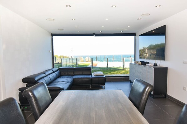 A modern living room with a large window, a black leather sofa, a dining table, and a flat-screen TV mounted on the wall.