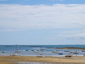 Ein Strand mit in flachem Wasser verankerten Booten, einem sandigen Ufer und einer in der Ferne liegenden felsigen Küstenlinie unter einer teils bewölkten Himmel.