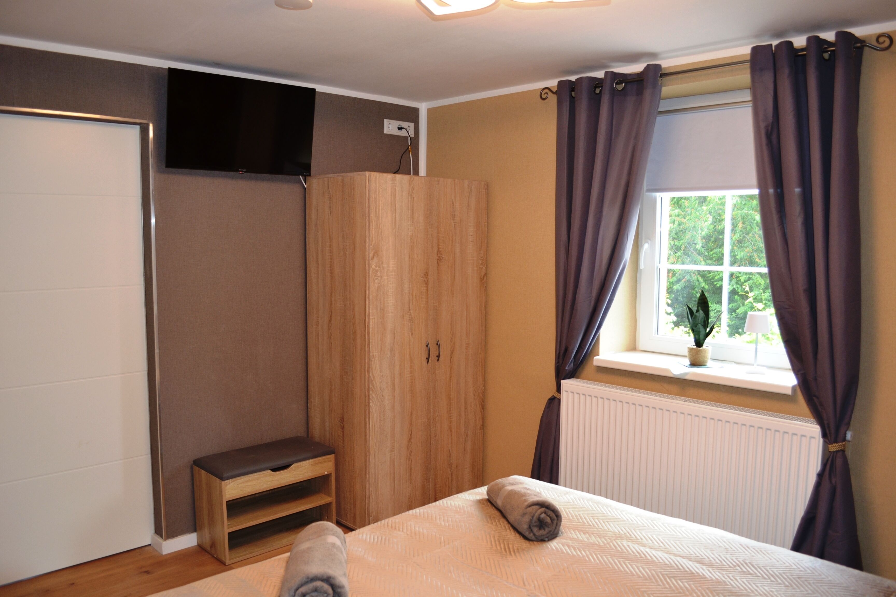 Une chambre à coucher avec un lit, une armoire en bois, un banc, une fenêtre avec des rideaux et un radiateur.