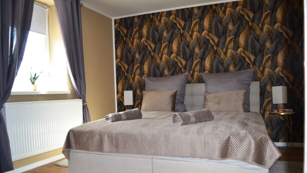 A bedroom with a bed, a window with curtains, a radiator, a wall with a leaf pattern, and a small table with a lamp. - Ferienwohnung Klüger - Vacation for the small family (Liebstadt)